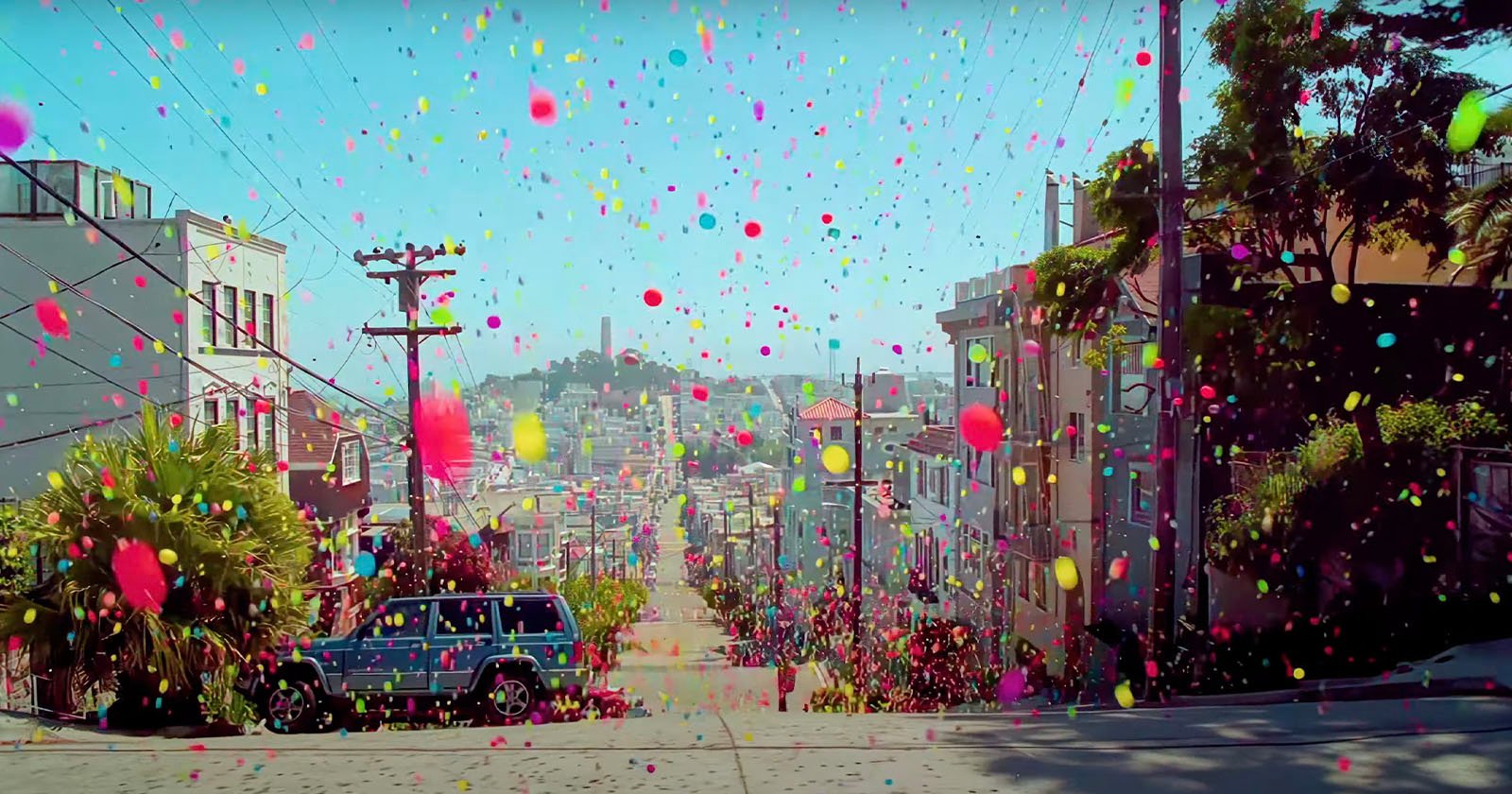 A steep urban street is filled with colorful, bouncing balls. The scene is vibrant, with various houses on either side. A parked SUV is visible on the left, and the sky is clear and blue in the background.