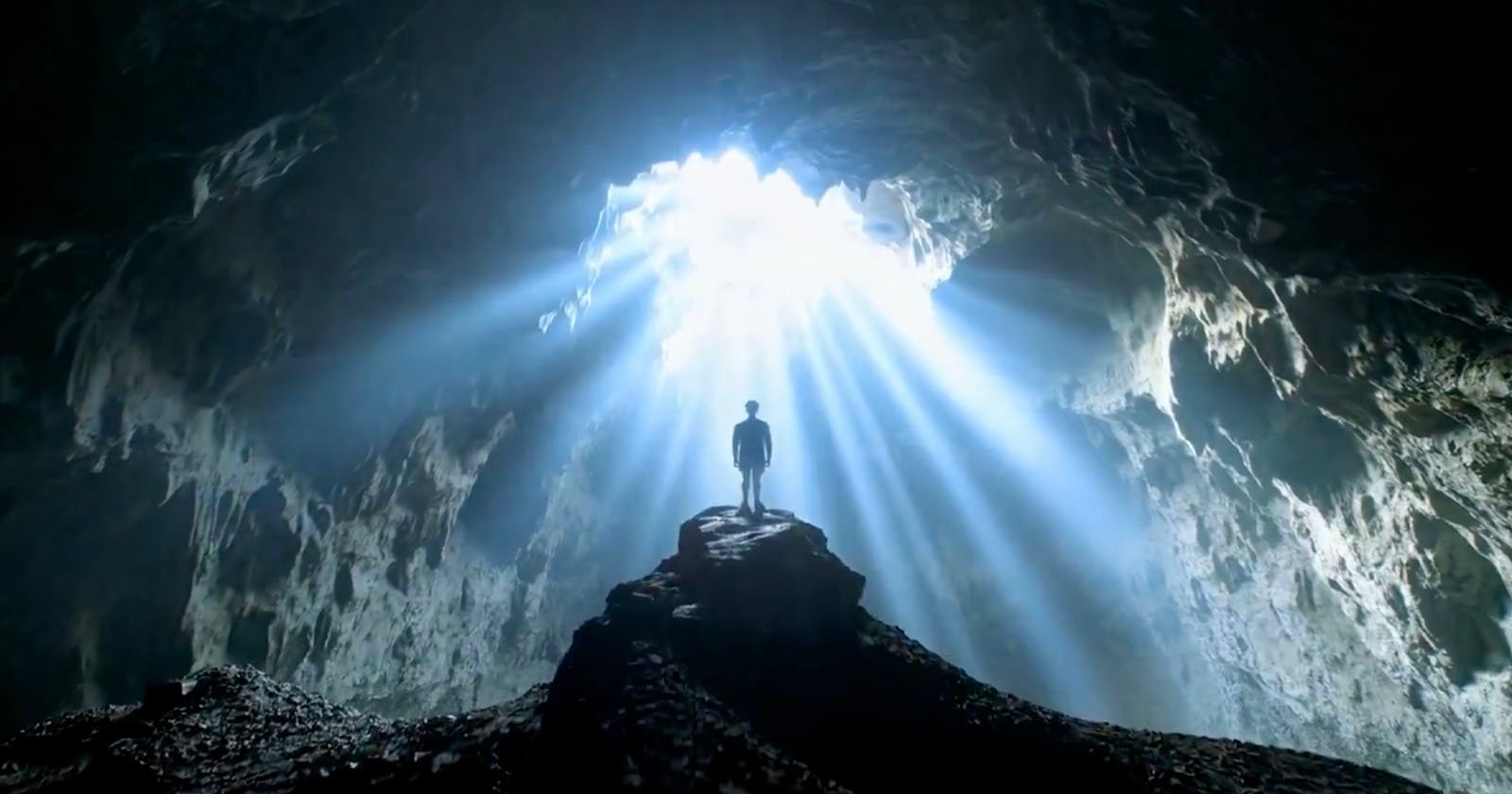 Silhouette of a person standing on a rock inside a cave, illuminated by beams of light streaming through an opening above. The scene is dramatic, highlighting the contrast between the dark cave interior and bright outside light.