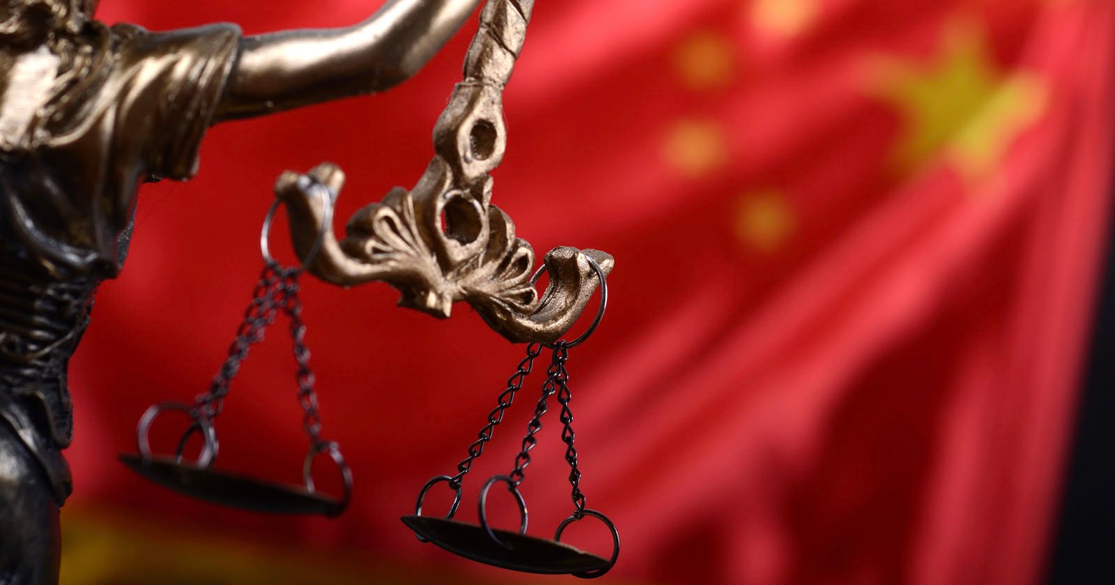 Close-up of a bronze Lady Justice statue holding scales, with the red flag of China featuring yellow stars blurred in the background.