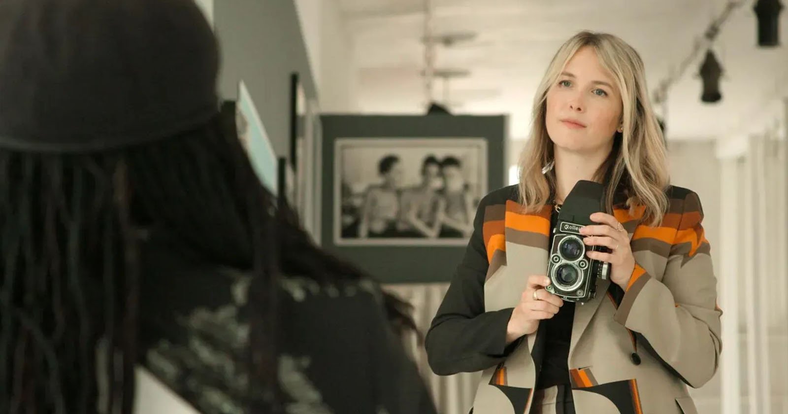 A woman with blonde hair holds a vintage camera and looks intently towards a person with long hair and a cap, visible from the back. They're standing in a bright room with photos on the wall.