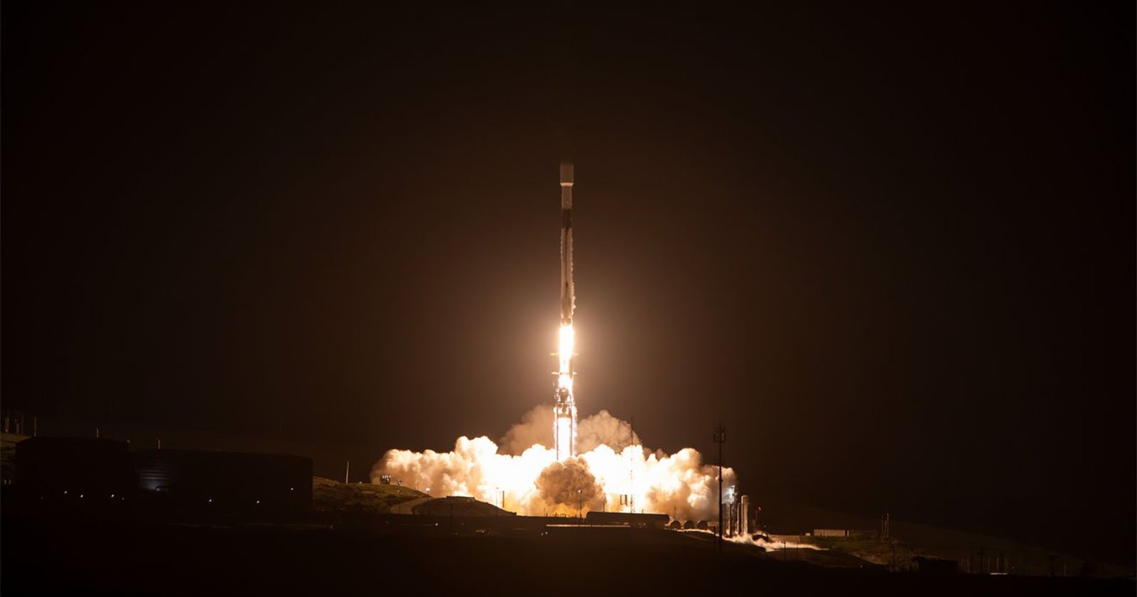 A rocket launches into the night sky, emitting bright flames and smoke from its engines. The dark background contrasts with the luminous glow of the rocket, highlighting its ascent from the ground.