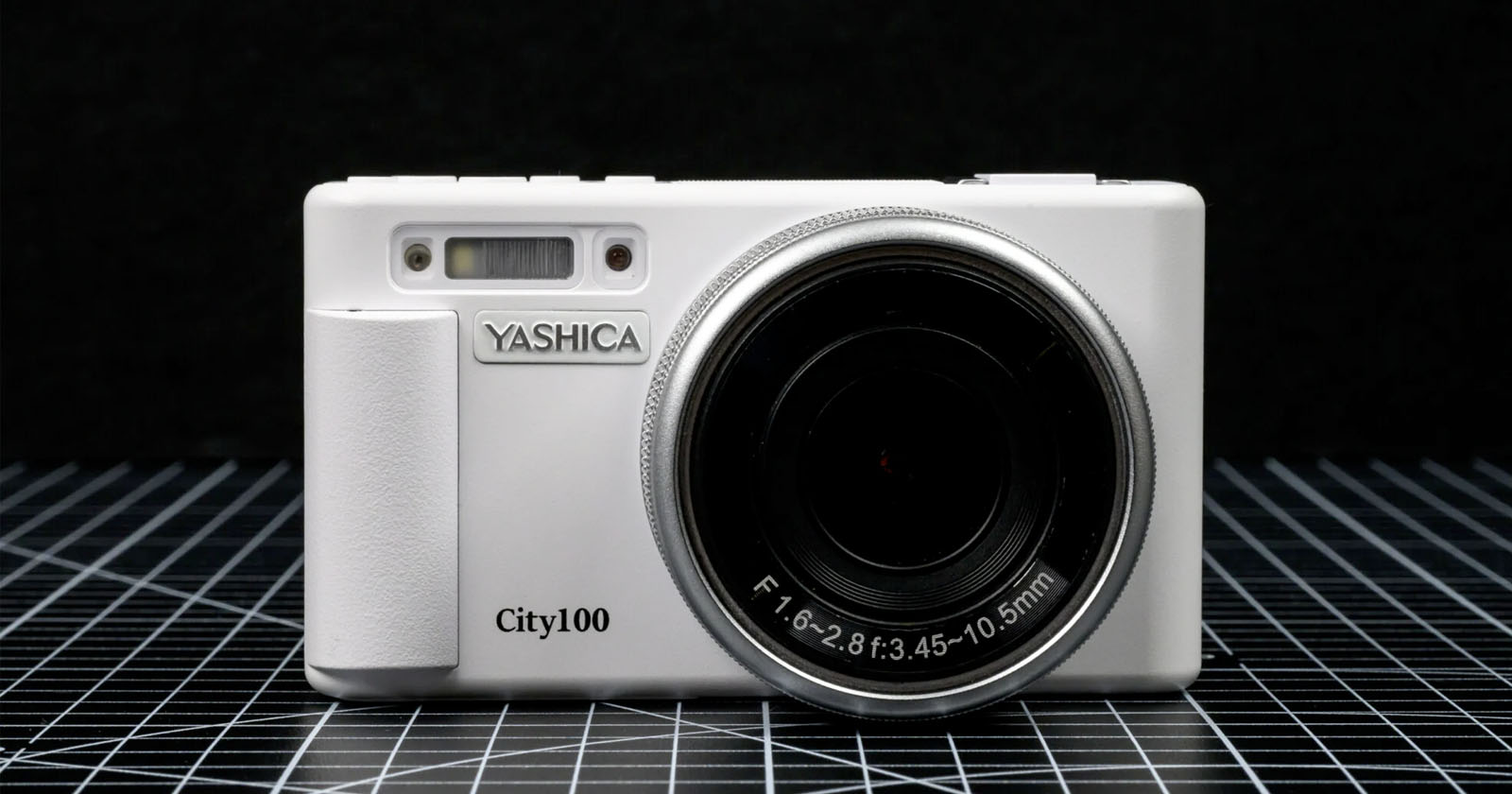 A white Yashica City100 digital camera is displayed upright on a grid-patterned surface against a black background, showing its lens and front details clearly.