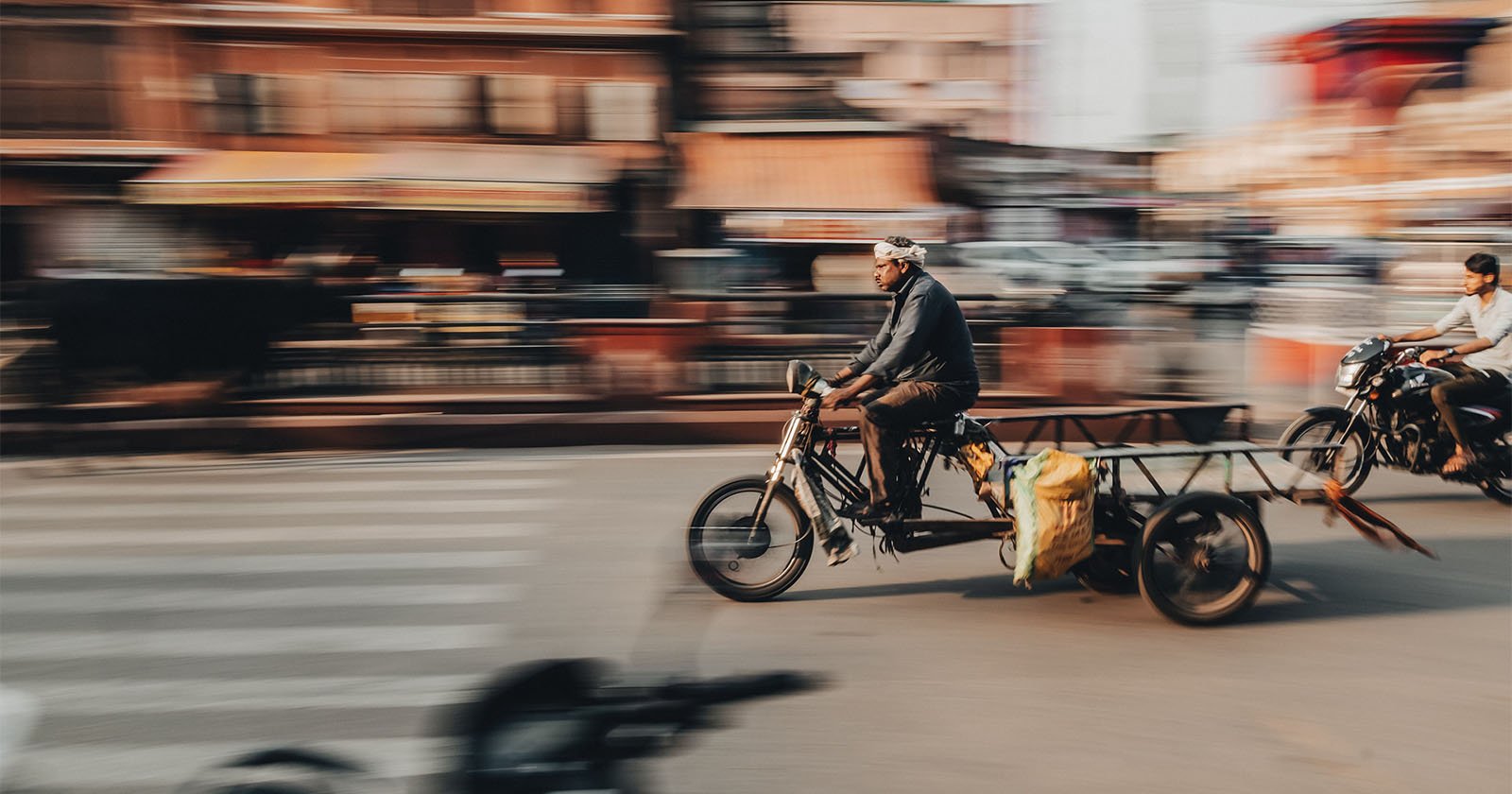 A man rides a motorbike with an attached cart down a busy street, motion blur showing speed; another motorcyclist follows, with buildings and pedestrians in the blurred background.