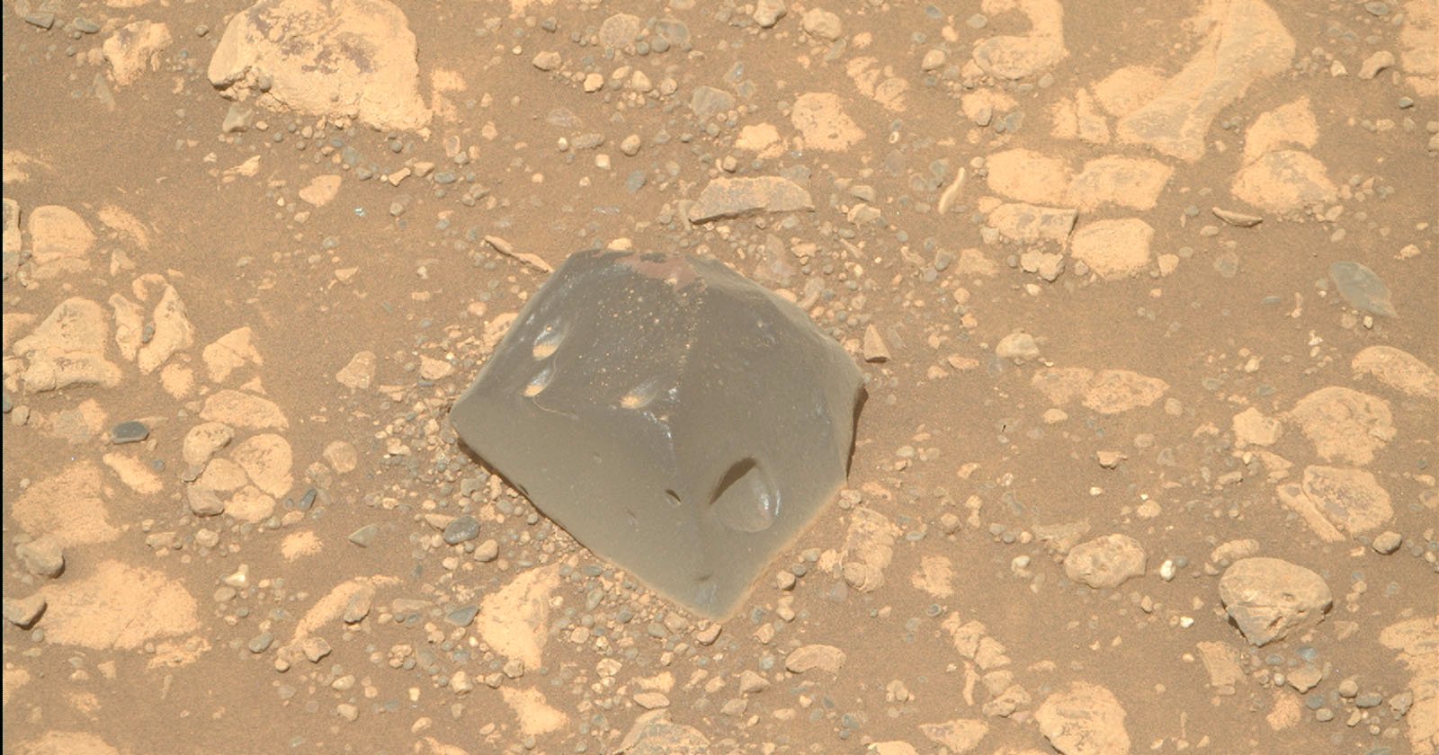A smooth, gray, glassy rock with angular edges sits among rough, beige, and dusty rocks and pebbles on a sandy surface.