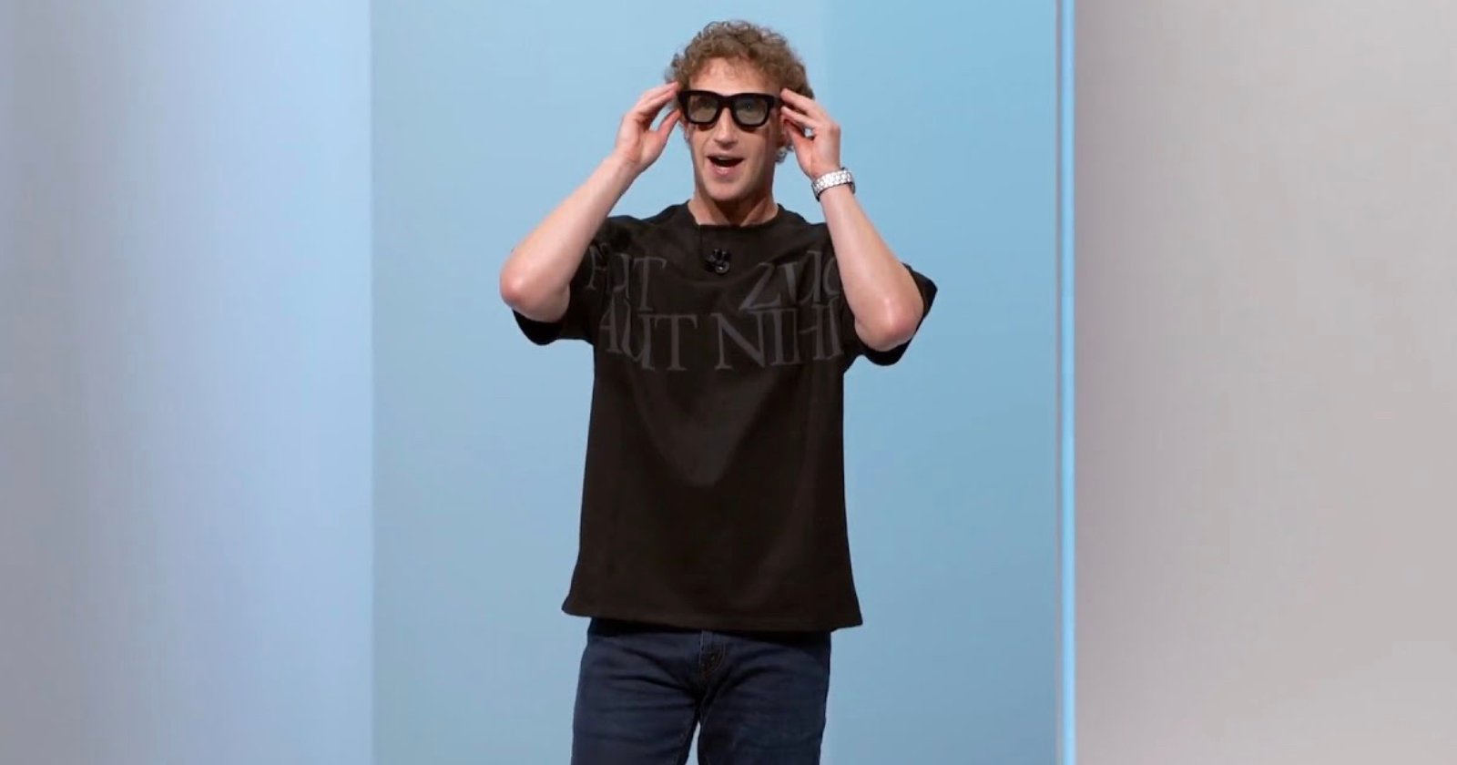 A person with curly hair wearing a black T-shirt, jeans, and large black glasses stands against a blue and white background, smiling with hands touching the glasses.